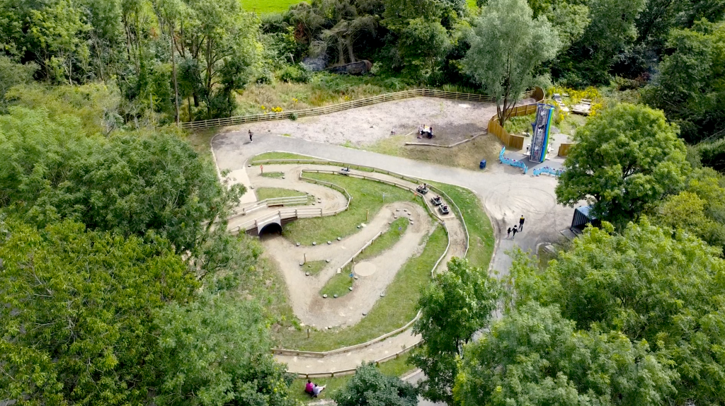 Drone shot of a looping off-road buggy track in a woodland clearing, with a tunnel, tyre-lined bends and a climbing tower nearby.
