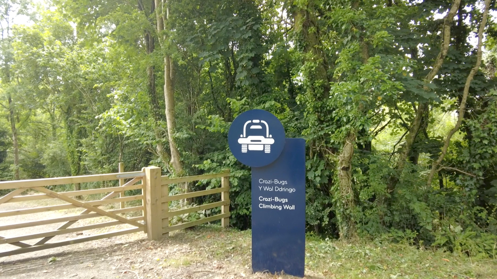 Blue Aqualand wayfinding sign with a buggy icon, marking the Crazi-Bugz and climbing wall area in bilingual Welsh and English, beside a wooden gate at a woodland entrance.